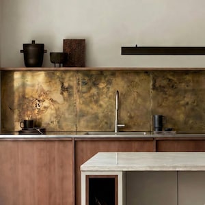 Pode incluir: Uma cozinha moderna com armários de madeira e um backsplash de latão. A bancada é de cor clara, com uma pia e torneira de aço inoxidável. Cerâmica preta decorativa e uma caneca preta estão na bancada.