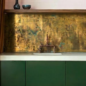 Può includere: Interno cucina con mobili bassi verde scuro e piano di lavoro bianco. Il paraschizzi è in metallo strutturato marrone dorato. Un rubinetto in rame è montato sul piano di lavoro. Vasi decorativi e una ciotola su una mensola di legno.
