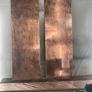 Two rectangular copper panels with a textured, aged appearance. The panels are displayed vertically, showcasing a rich, reddish-brown colour with variations in tone. The surface has a mottled effect, suggesting a natural or weathered finish.