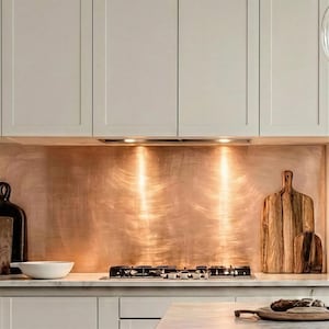 May include: A modern kitchen with light gray cabinets and a copper backsplash. Two wooden cutting boards and a white bowl sit on the countertop. A gas cooktop is visible, and the scene is illuminated by recessed lighting.