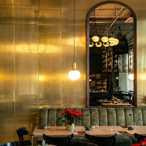 May include: A restaurant interior featuring a brass-colored wall, a large arched mirror, and a green velvet booth. Hanging from the ceiling are several globe-shaped lights. A table is set with glasses and a poinsettia plant.
