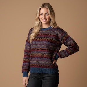 May include: A woman wearing a patterned jumper with a blue crew neck and cuffs. The jumper features a geometric design in shades of red, blue, and gold. The background is a solid tan colour.