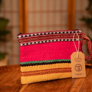May include: A pink, yellow, green, and black woven clutch bag with a brown tag that reads "Handcrafted treasures from the Andes." The tag also includes a logo with a stylized bird and the text "www.andescraft.com Made in Peru."