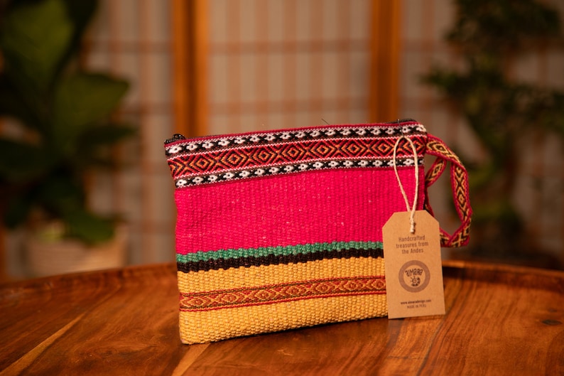 May include: A pink, yellow, green, and black woven clutch bag with a brown tag that reads "Handcrafted treasures from the Andes." The tag also includes a logo with a stylized bird and the text "www.andescraft.com Made in Peru."