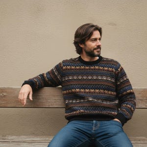 Könnte beinhalten: Ein gemusterter Pullover in Braun-, Blau- und Schwarztönen. Der Pullover hat einen Rundhalsausschnitt und lange Ärmel. Die Person trägt eine blaue Jeans und sitzt auf einer Holzbank.