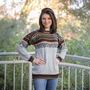 May include: A woman wearing a grey and brown patterned jumper with long sleeves. The jumper has a geometric design with green, brown, and white accents.