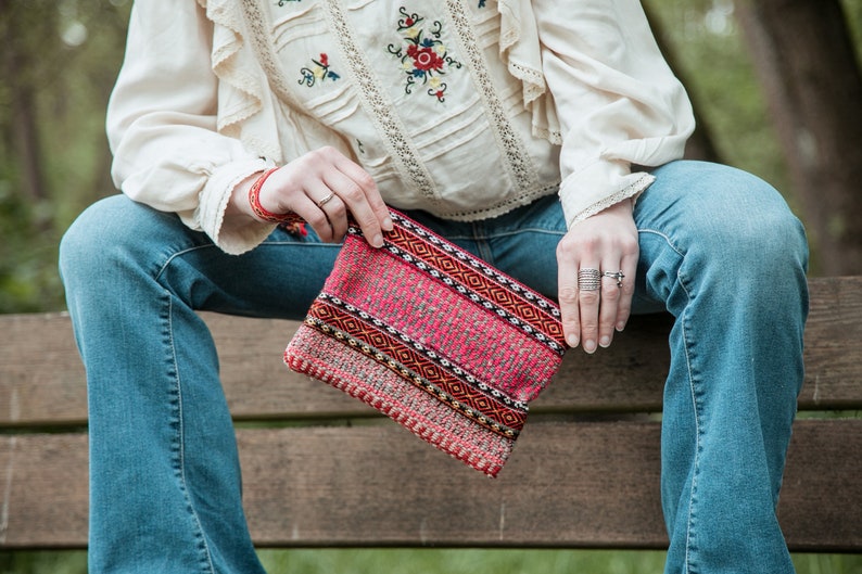 May include: A woman is sitting on a wooden bench holding a pink and red woven clutch with geometric patterns.