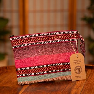 May include: A pink and blue woven clutch bag with a geometric pattern and a brown tag that reads "Handcrafted treasures from the Andes".