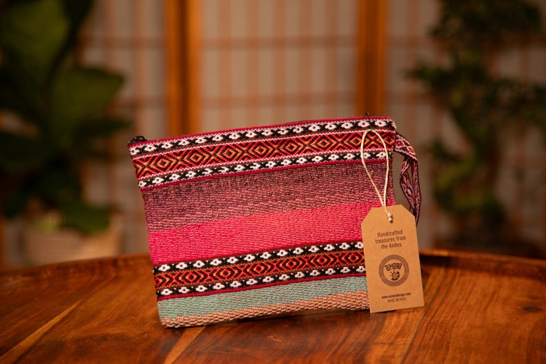 May include: A pink and blue woven clutch bag with a geometric pattern and a brown tag that reads "Handcrafted treasures from the Andes".