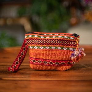 Könnte beinhalten: Kleine, gewebte orangefarbene Tasche mit schwarzem Reißverschluss, rotem Riemen und dekorativem Bommel. Die Tasche weist komplizierte geometrische Muster in Rot, Weiß, Grün und Schwarz auf. Der Riemen ist ca. 15 cm lang.