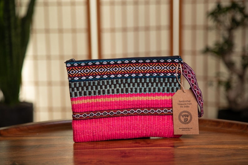May include: A pink woven clutch with a colorful geometric pattern and a brown tag that reads "Handcrafted treasures from the Andes".
