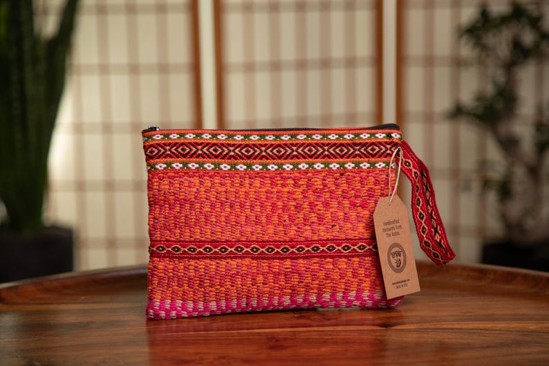 May include: A red, orange, and pink woven clutch bag with a black zipper and a brown tag that says "Handmade from The Andes".