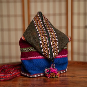 Może przedstawiać: Kolorowa, tkana torba z zamkiem błyskawicznym i frędzlem. Torba jest wykonana z różnych tkanin, w tym zielonej, czerwonej, niebieskiej i białej. Torba leży na drewnianym stole.