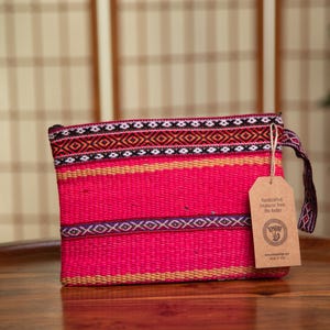 May include: A pink woven clutch bag with a black, white, and orange geometric pattern. The bag has a brown tag that says "Handcrafted Treasures from The Andes".