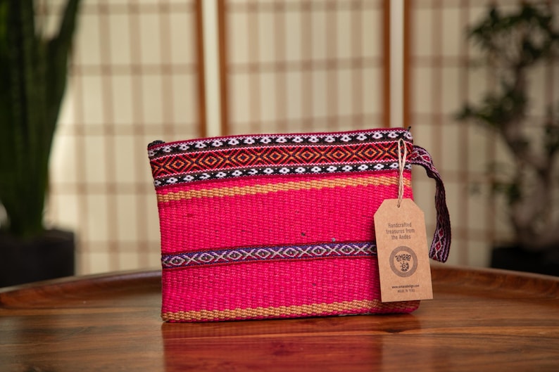 May include: A pink woven clutch bag with a black, white, and orange geometric pattern. The bag has a brown tag that says "Handcrafted Treasures from The Andes".