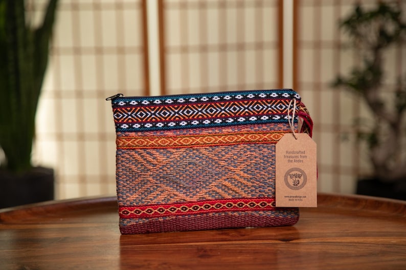 May include: A colorful woven clutch bag with a zipper closure. The bag has a brown paper tag attached with the text "Handcrafted Treasures from the Andes" and a logo.