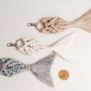 May include: Three macrame fish tail keychains in gray, white, and beige. Each keychain has a silver ring and a tassel. The keychains are arranged in a row on a white surface. A small wooden tag with the text "Made with Love" is visible in the bottom right corner of the image.