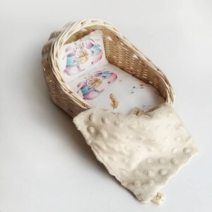 May include: A white wicker doll cradle with a white and pink floral patterned sheet and a white pillow with a print of two bunnies wearing sweaters. A cream colored blanket with white polka dots is draped over the cradle.