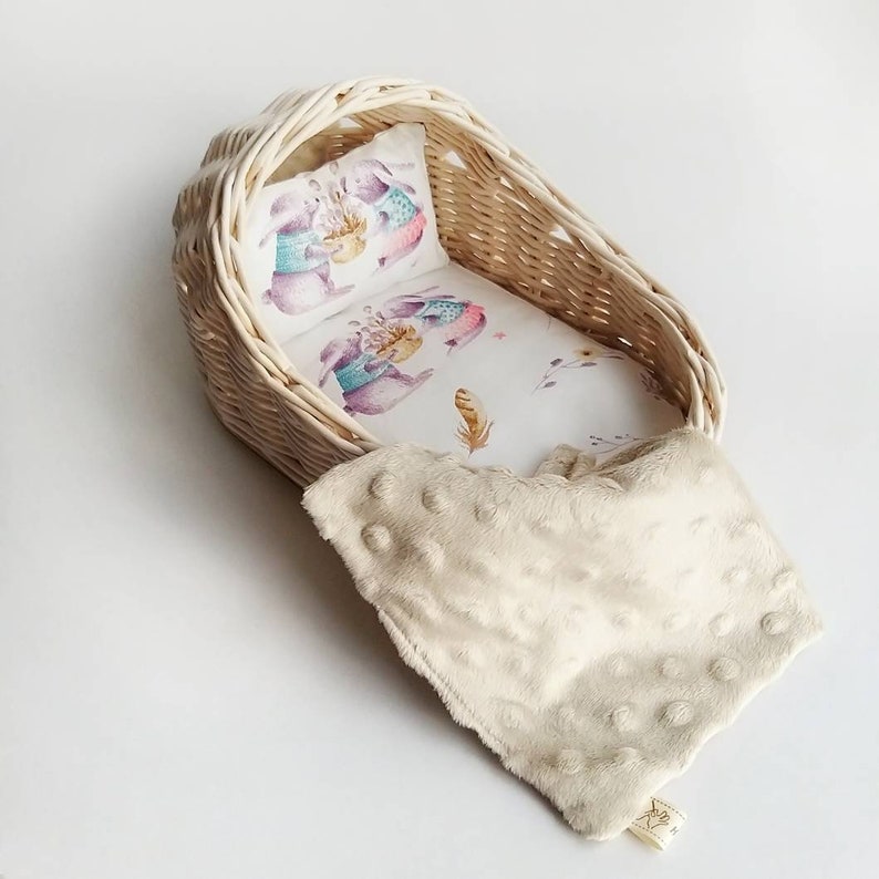 May include: A white wicker doll cradle with a white and pink floral patterned sheet and a white pillow with a print of two bunnies wearing sweaters. A cream colored blanket with white polka dots is draped over the cradle.