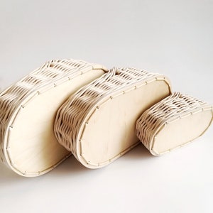 May include: Three oval-shaped wicker baskets with wooden bottoms, nested together in decreasing size. The baskets are a light cream color.