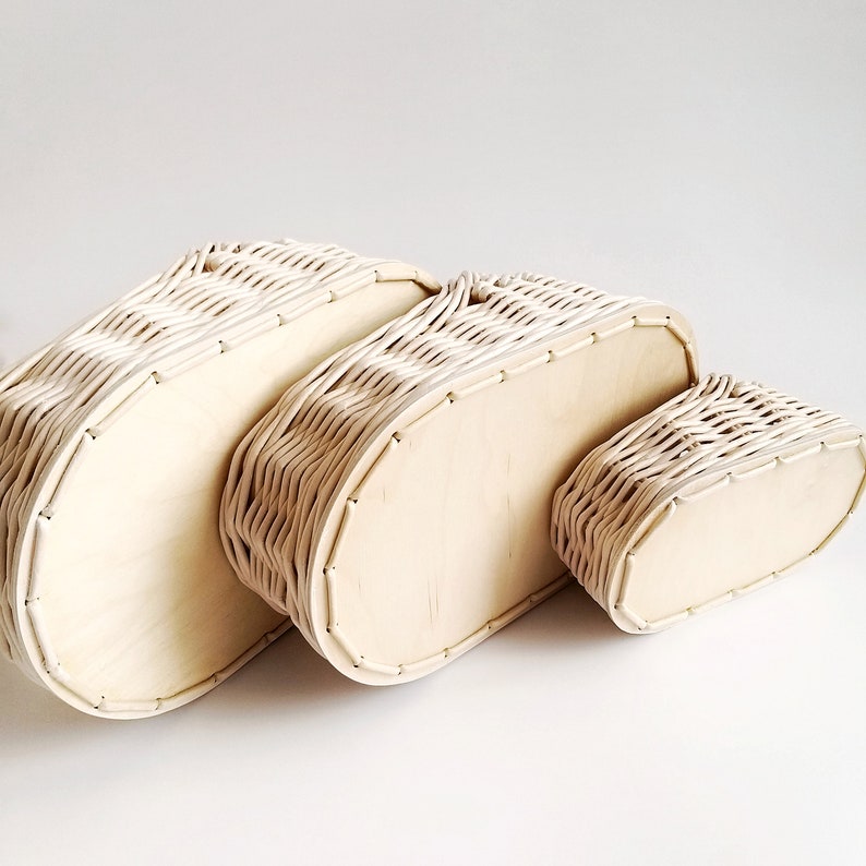 May include: Three oval-shaped wicker baskets with wooden bottoms, nested together in decreasing size. The baskets are a light cream color.