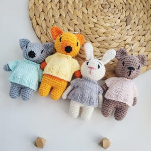 Puede incluir: Cuatro llaveros de animales tejidos a mano: un oso gris, un oso amarillo, un conejo blanco y un oso marrón. Cada peluche lleva un pequeño suéter de diferente color pastel. Los llaveros están colocados frente a un salvamanteles circular tejido.