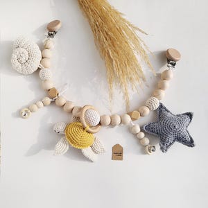 May include: A baby pacifier clip with wooden beads, a crocheted star, a crocheted turtle with a yellow shell, and a crocheted seashell. The clip has two wooden clips and a wooden ring.