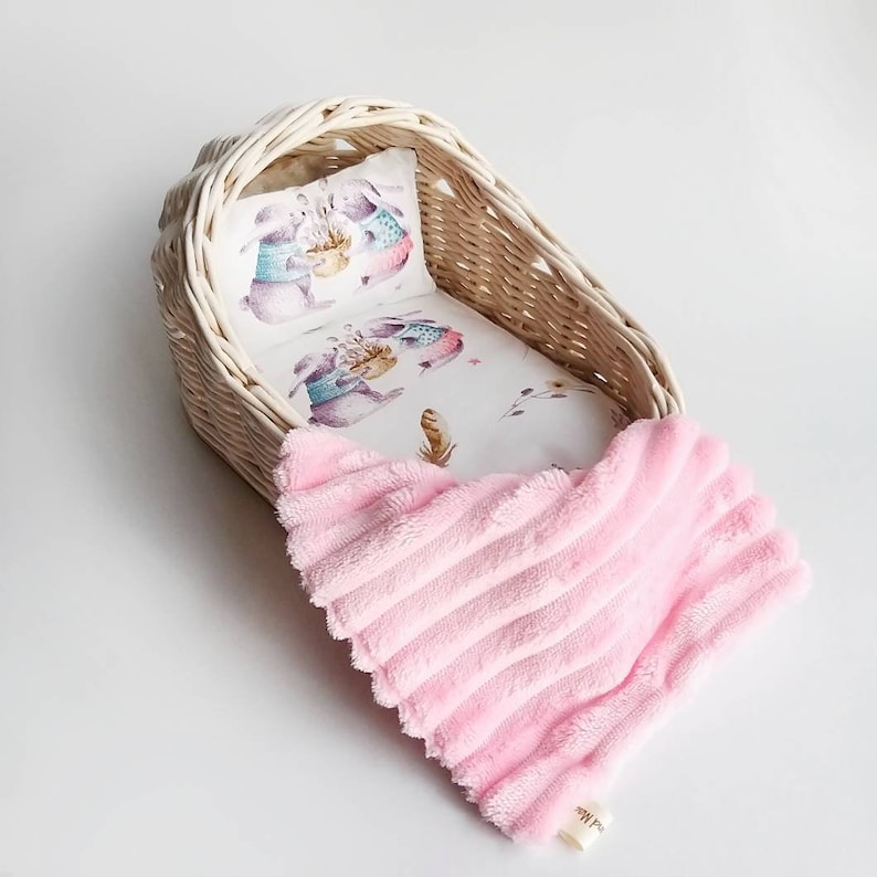 May include: A white wicker doll cradle with a white and pink floral patterned sheet and a pink plush blanket. The sheet has a design of two bunnies wearing sweaters.