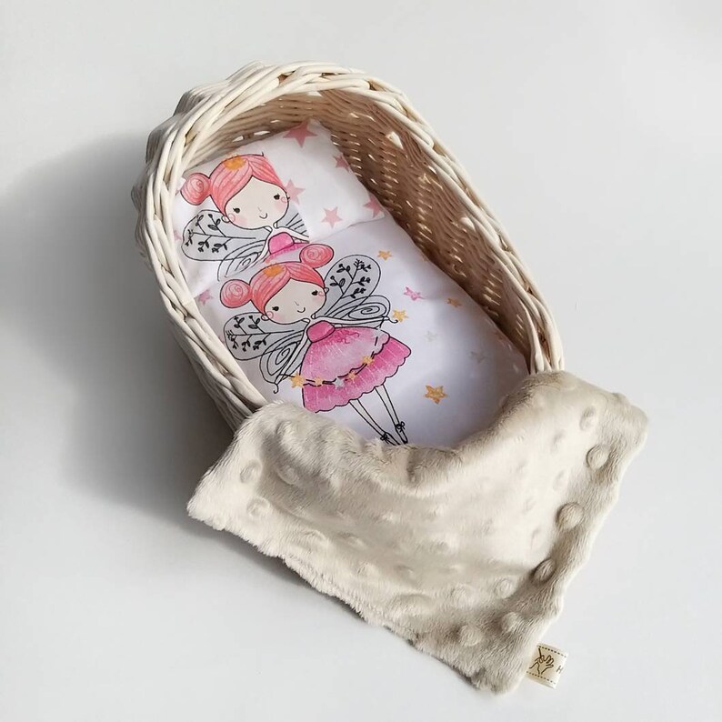May include: A white wicker doll crib with a white and pink fairy-themed bedding set. The crib is filled with a white plush blanket with small raised dots.