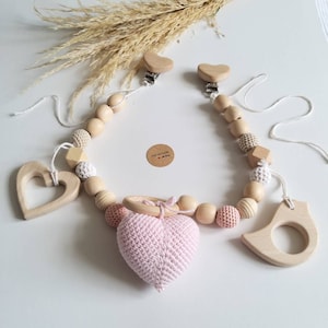 May include: A baby pram chain featuring natural wooden beads, crochet-covered beads in beige and pink, two heart-shaped wooden clips, a wooden heart, a wooden bird shape, and a central pink crocheted heart. A small round tag reads Handmade with love.