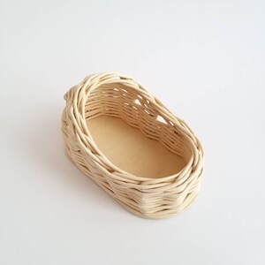 May include: A small, oval-shaped wicker basket with a light beige color. The basket has a woven design and a flat, wooden base.