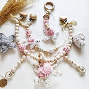 May include: A collection of handmade baby accessories, including pacifier clips and a wooden teether. The clips feature wooden beads, crocheted turtle shapes in white and pink, and wooden clips. A crocheted star and a seashell are also visible.