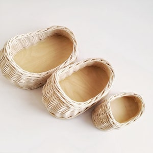 May include: Three white wicker baskets of varying sizes, each with a wooden base. The baskets are arranged in a decreasing size order from left to right.