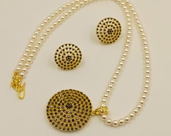 Puede incluir: Un conjunto de joyería compuesto por un collar y dos pendientes. El collar presenta una hilera de perlas blancas con un gran colgante circular dorado incrustado con pequeñas piedras negras. Los pendientes a juego son circulares y dorados.