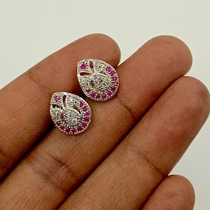 Könnte beinhalten: Ein Paar tropfenförmige Ohrstecker mit einer zentralen Ansammlung klarer Steine, umgeben von einem Rand aus leuchtend rosa Steinen in einem Metallrahmen. Das Design weist einen kleinen offenen Bereich innerhalb der Tropfenform auf.