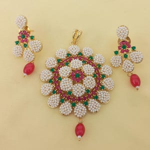 May include: A gold-coloured jewellery set comprising a pendant and matching earrings. The pendant and earrings are decorated with white pearl-like beads, red and green gemstones. Each piece has a red teardrop-shaped bead suspended from the bottom.