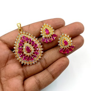 Puede incluir: Un conjunto de joyería en tono dorado que consta de un gran colgante en forma de pera y dos pendientes a juego. Cada pieza presenta una piedra rosa central en forma de lágrima rodeada de piedras rosas más pequeñas y cristales blancos.