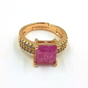 Op de afbeelding: Een goudkleurige verstelbare ring met een grote, vierkante, getextureerde felroze middensteen die door vier pootjes wordt vastgehouden. De ringband is verdeeld in twee rijen, elk bezet met kleine, heldere, sprankelende steentjes aan de zijkanten.