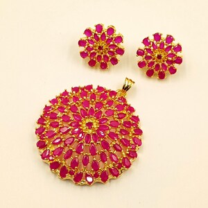 May include: A gold-toned jewelry set consisting of a large circular pendant and two matching circular stud earrings. Each piece features a floral-inspired arrangement of numerous vibrant pink, pear-shaped stones set in a bright gold metal frame.