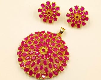 Può includere: Un set di gioielli color oro composto da un grande ciondolo circolare e due orecchini a lobo abbinati. Ogni pezzo presenta un motivo floreale con numerose pietre rosa brillante a forma di goccia incastonate in una montatura in metallo dorato.