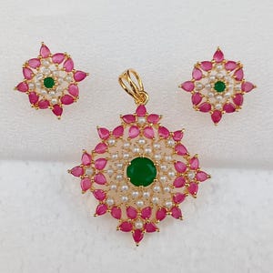 Puede incluir: Un conjunto de joyas dorado con un colgante y pendientes a juego. El colgante y los pendientes tienen un diseño floral con acentos rosas en forma de pétalo, pequeñas perlas blancas y una piedra verde central. El conjunto se muestra sobre una superficie blanca.