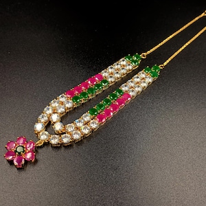 May include: A gold-tone necklace featuring two parallel strands of round-cut white, pink, and green stones. The necklace terminates in a flower-shaped pendant composed of six pink petals surrounding a single central green stone.