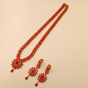 May include: A gold-toned necklace and matching earring set featuring vibrant red, teardrop-shaped stones arranged in a floral pattern. The necklace has a long chain of linked red stones, and each earring displays a central red stone surrounded by petals.