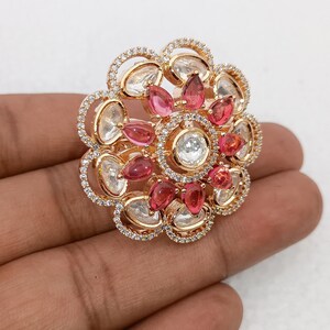 Puede incluir: Un anillo dorado con un diseño floral. El anillo presenta una piedra central transparente rodeada de piedras rosas y transparentes. Los bordes exteriores del anillo están adornados con pequeñas piedras transparentes, creando un efecto brillante. El anillo se sostiene en una mano.