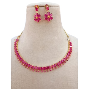 Puede incluir: Un conjunto de collar y pendientes dorados. El collar presenta una serie de piedras en forma de lágrima, de color rubí. Los pendientes tienen forma de flor, con pétalos de color rubí y un centro dorado. Las joyas se exhiben en un maniquí.