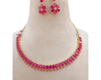 May include: A gold-toned necklace and earring set. The necklace features a series of teardrop-shaped, ruby-colored stones. The earrings are flower-shaped, with ruby-colored petals and a gold-colored center. The jewelry is displayed on a mannequin.
