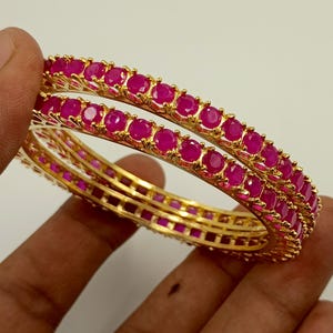May include: Two gold-toned bangles featuring a continuous row of round, vibrant pink stones set in a prong setting. The metal bands have a polished finish, and the stones are uniform in size and colour, creating a circular design for the wrist.
