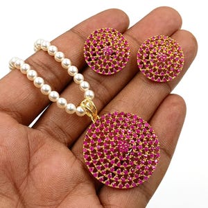 Puede incluir: Un conjunto de joyería compuesto por un collar y dos pendientes. El collar presenta una hilera de perlas blancas con un colgante circular de tono dorado incrustado con pequeñas piedras de color rosa vibrante. Los pendientes a juego son circulares.