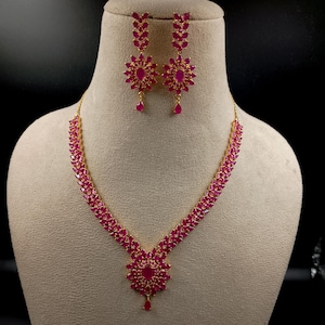 Puede incluir: Un collar dorado y un par de pendientes a juego, con piedras de color fucsia intenso en talla marquesa dispuestas en patrones florales y de hojas, con una piedra ovalada central y un pequeño detalle en forma de lágrima.