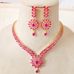Puede incluir: Un conjunto de joyas dorado con un collar y pendientes a juego. El collar y los pendientes están adornados con piedras preciosas de color rosa vibrante dispuestas en un patrón floral. El conjunto incluye un colgante y pendientes colgantes.